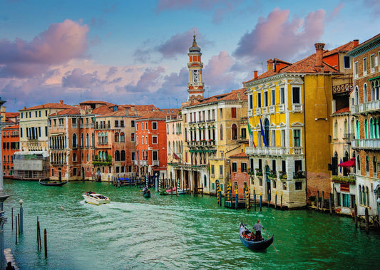 Venice Canal sunset photo in Italy. The water is a pretty blue green and there are people in canoes on the water. The buildings are painted light yellow, cream color and orange. This print is great for any room of the home.