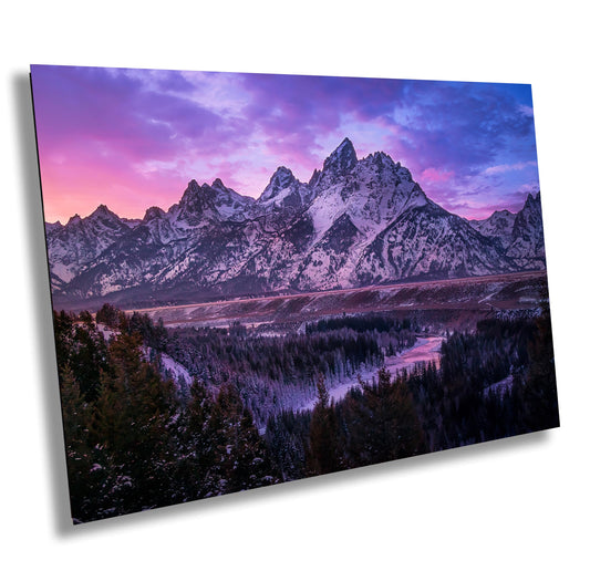 Snow-dusted peaks rise majestically against a sky painted in soft pink and purple hues. Below, a winding river meanders through a dark forest, its snowy banks glowing faintly in the twilight, creating a serene and timeless winter landscape.